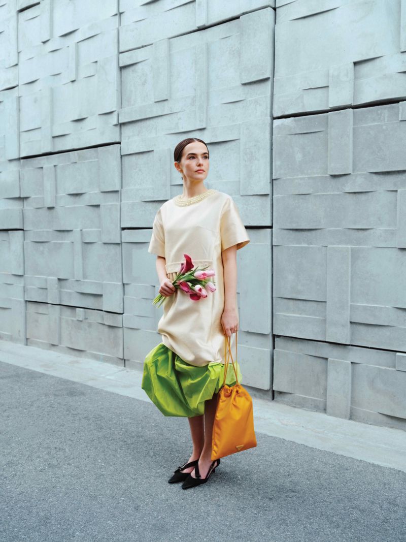 Woman in a cream-colored dress stands on an asphalt surface holding pink tulips and a drawstring bag in front of a block wall.