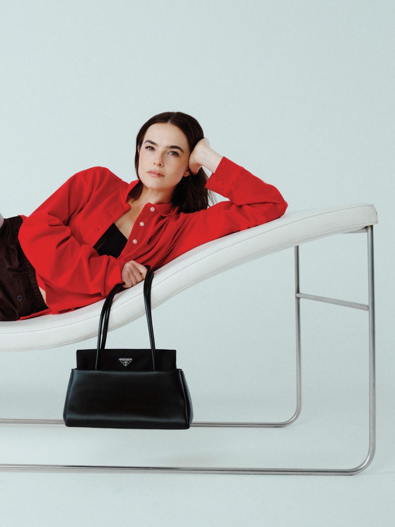 A woman poses on a white chaise lounge next to a black Prada handbag against a white backdrop in a studio fashion shot.