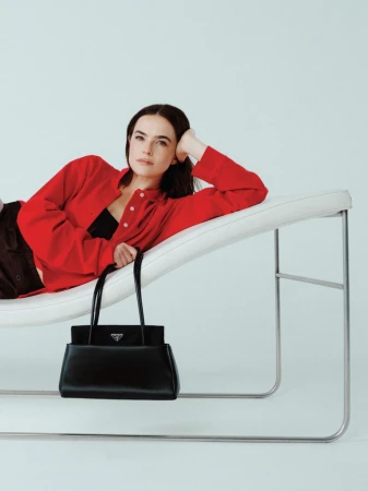 A woman poses on a white chaise lounge next to a black Prada handbag against a white backdrop in a studio fashion shot.
