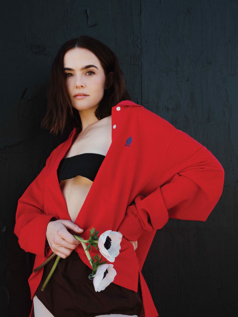 A fair-skinned woman in a red jacket holds white flowers in front of a dark background, captured in a studio-style portrait.