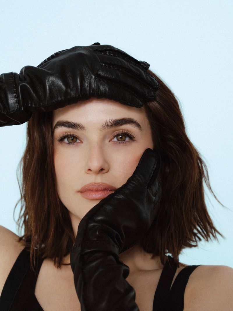 Close-up of a brunette adult woman with her face framed by black leather gloves, set against a soft light blue background.