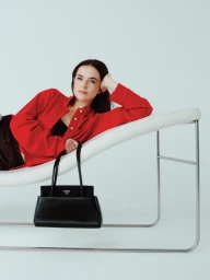 Zoey Deutch reclining on a white lounge chair wearing a red button-up shirt and holding a black handbag.
