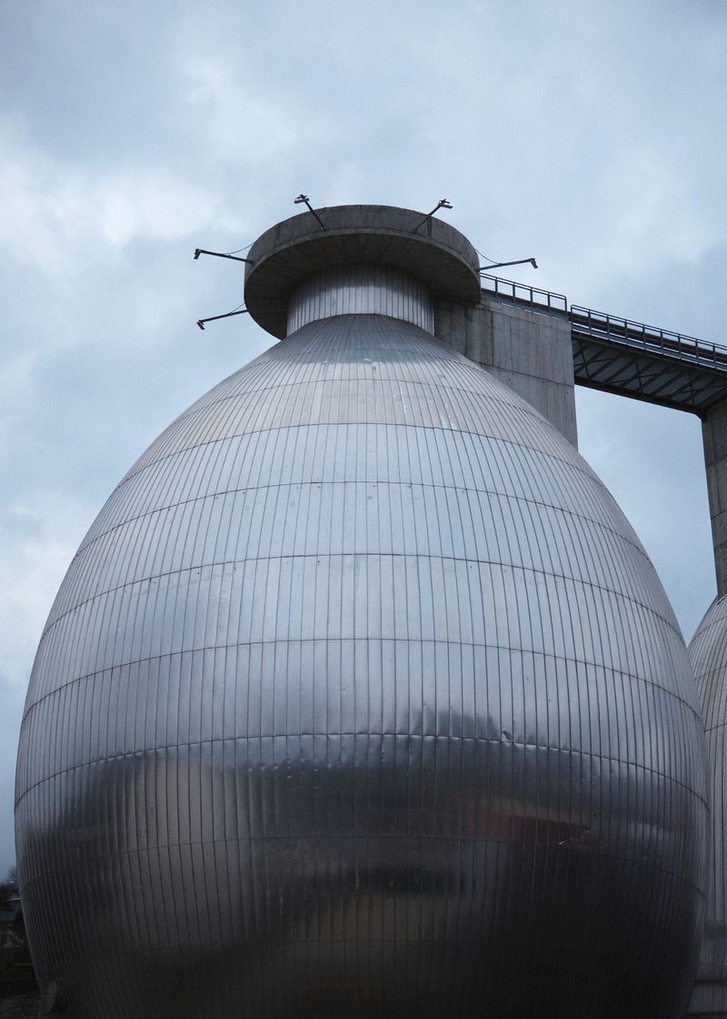 Industrial metal digester tanks with rounded tops, reflecting the sky, alongside a gray concrete support structure under an overcast sky.