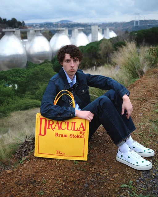 Adult male poses with a 'Dracula by Bram Stoker' tote bag outdoors against a background of large metal tanks and forest.