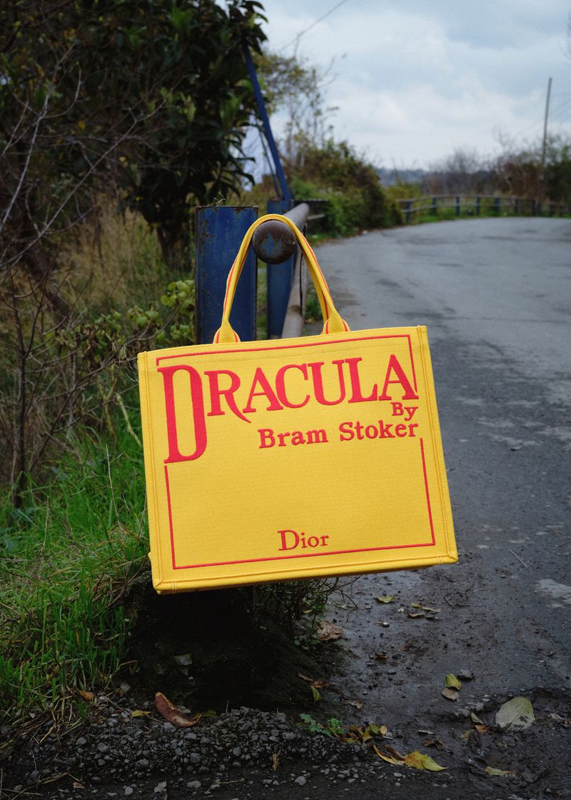 Yellow tote bag with 'Dracula' text hanging from a blue metal bar, photographed outdoors with a blurred background of trees and sky.