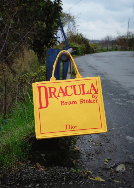 Yellow tote bag with 'Dracula' text hanging from a blue metal bar, photographed outdoors with a blurred background of trees and sky.