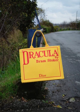 Yellow tote bag with 'Dracula' text hanging from a blue metal bar, photographed outdoors with a blurred background of trees and sky.