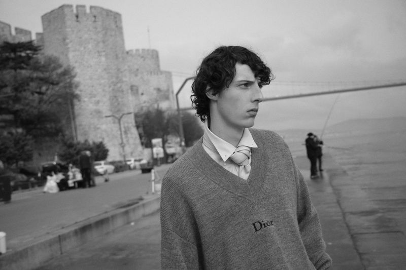 Portrait of a young adult man in a Dior sweater standing near a waterside, with an old stone fortress and bridge in the background.