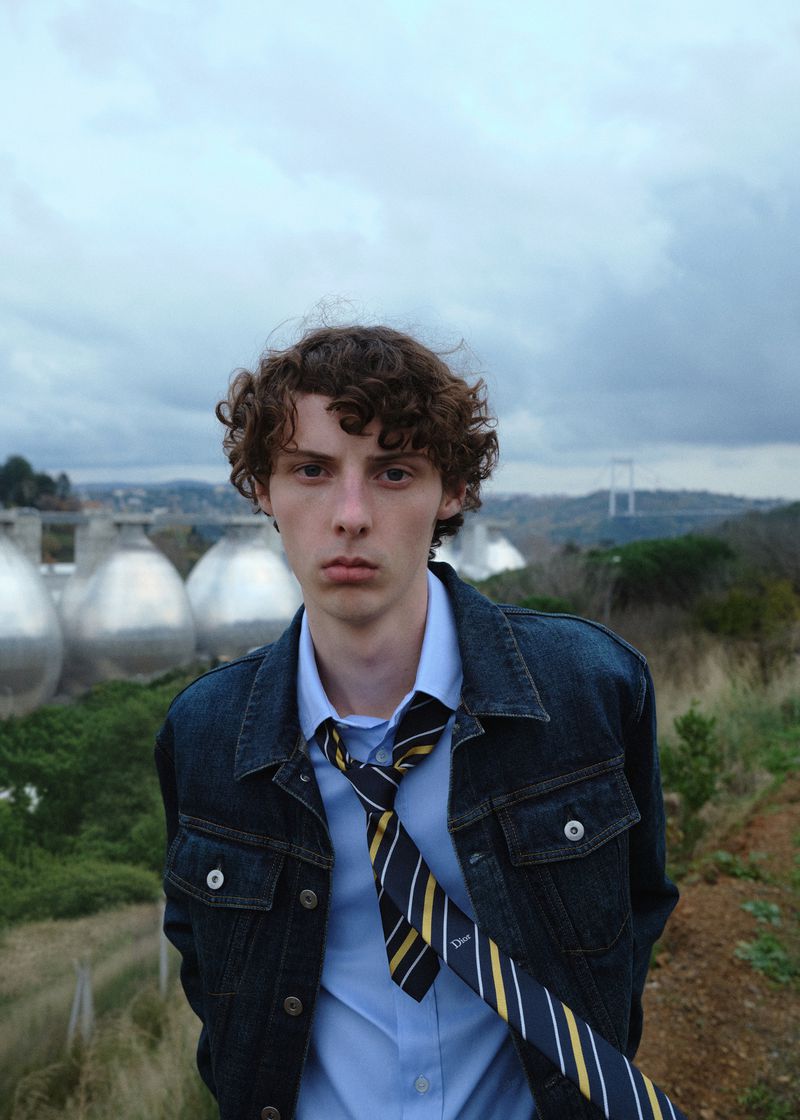 Young man with curly hair in denim jacket and tie stands outdoors near industrial tanks, under an overcast sky with distant bridge.