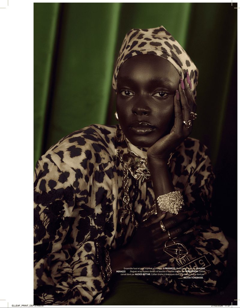 Elegant dark-skinned woman in leopard-print headscarf and jacket, adorned with jewelry, poses against a green draped backdrop.