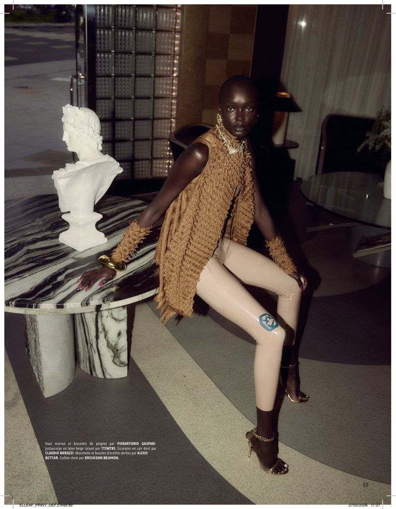 Fashion editorial shot of a Black female model in a woven vest and shiny leggings, posing near a marble table with a bust in an indoor setting.