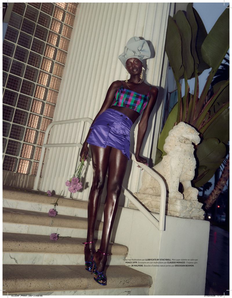 Fashion editorial shot of a Black female model in a purple mini skirt and plaid crop top, posing on outdoor stairs with flowers.