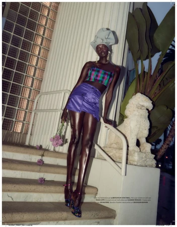 Fashion editorial shot of a Black female model in a purple mini skirt and plaid crop top, posing on outdoor stairs with flowers.