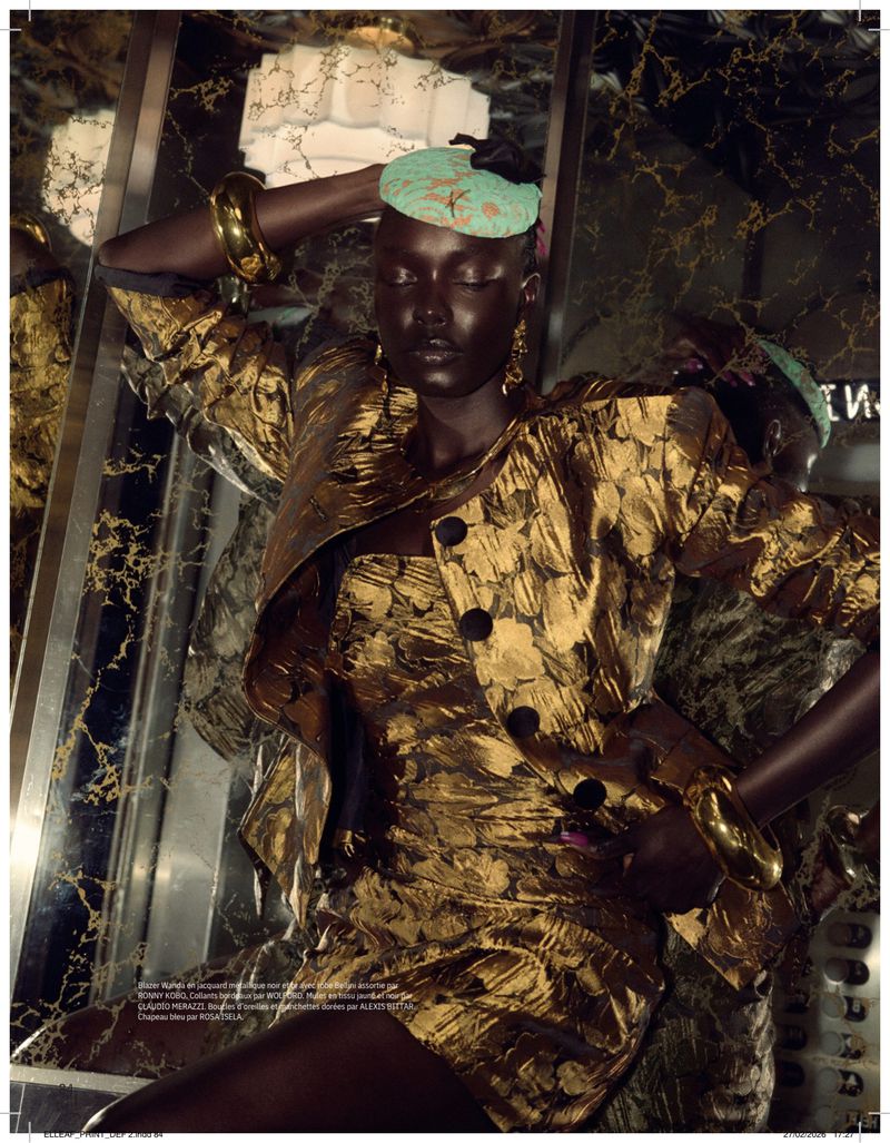 Woman in gold jacket and short skirt poses against marble wall. She wears a hat, gold jewelry, and has dark skin and closed eyes.
