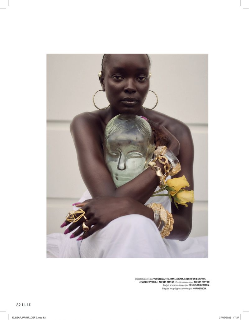 A dark-skinned woman in white clothing and jewelry holds a clear glass head with yellow roses, set against a neutral wall backdrop.