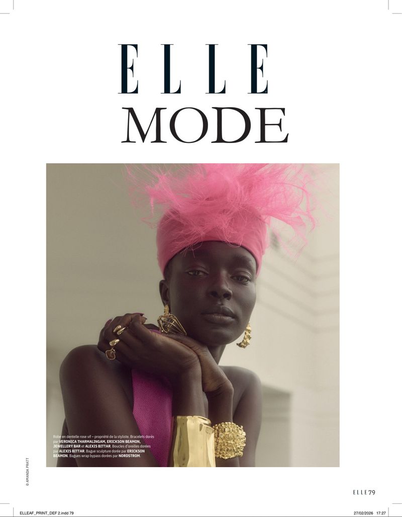 Close-up portrait of a dark-skinned woman wearing a pink feathered hat and gold jewelry, photographed indoors with neutral-toned walls.