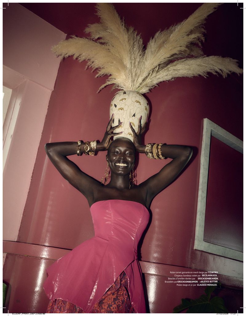 Elegant woman in pink strapless dress with jewelry, holding vase of pampas grass on her head against a pink wall indoors.