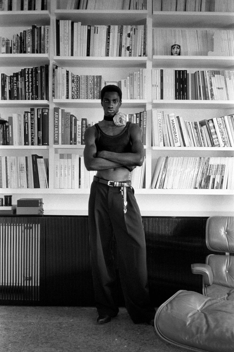 Black and white portrait of an African American adult male with crossed arms, large trousers, in front of bookshelf and chair.