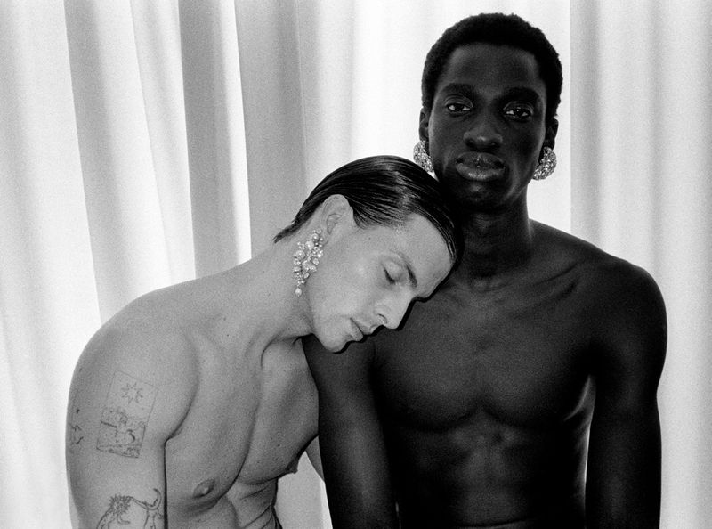 Monochrome studio shot featuring two shirtless adult men with dark and light skin tones, adorned with large, ornamental earrings, against a draped backdrop.