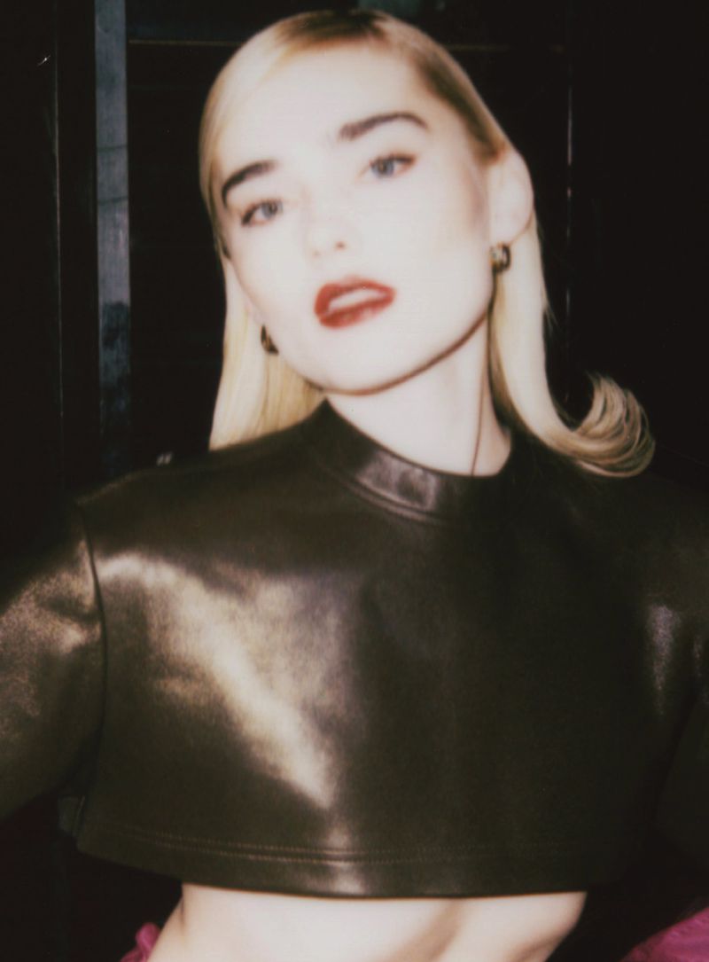 Portrait of a woman with blonde hair wearing a brown leather crop top, posing in a dark, indoor environment with soft, diffused lighting.