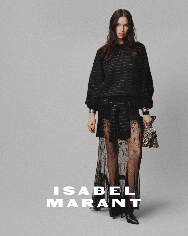 Fashion shot of a brunette adult female model wearing a black sweater and see-through skirt, accessorized with bracelets and a handbag, posing in a photo studio.