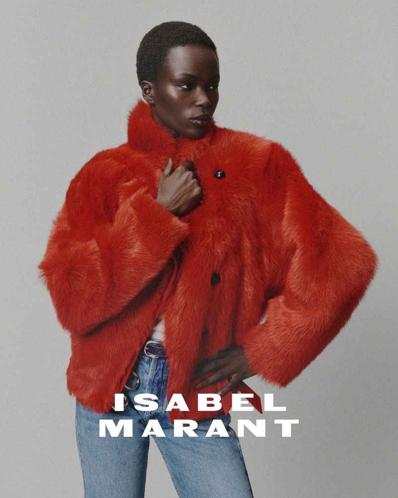 Fashion shot of a woman in a red faux fur coat and blue jeans against a gray backdrop, posing for a portrait, promoting clothing brand.