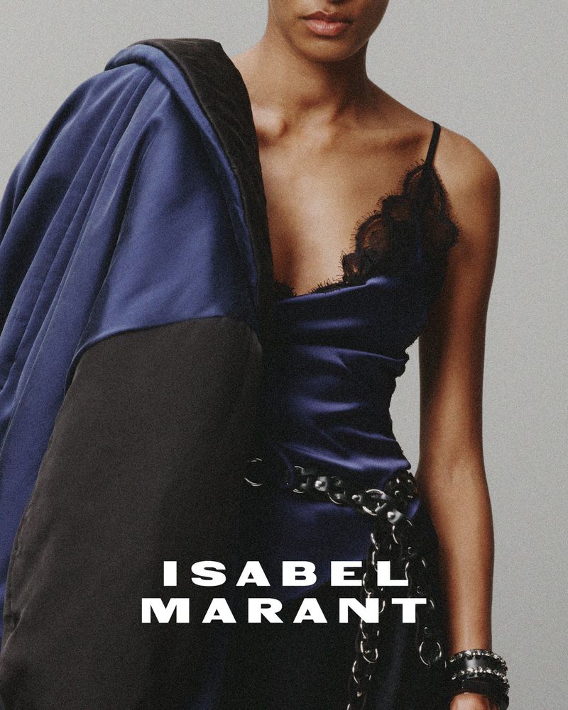 Fashion shot: adult female model poses in blue slip dress with black lace, paired with a draped jacket, against a grey background.