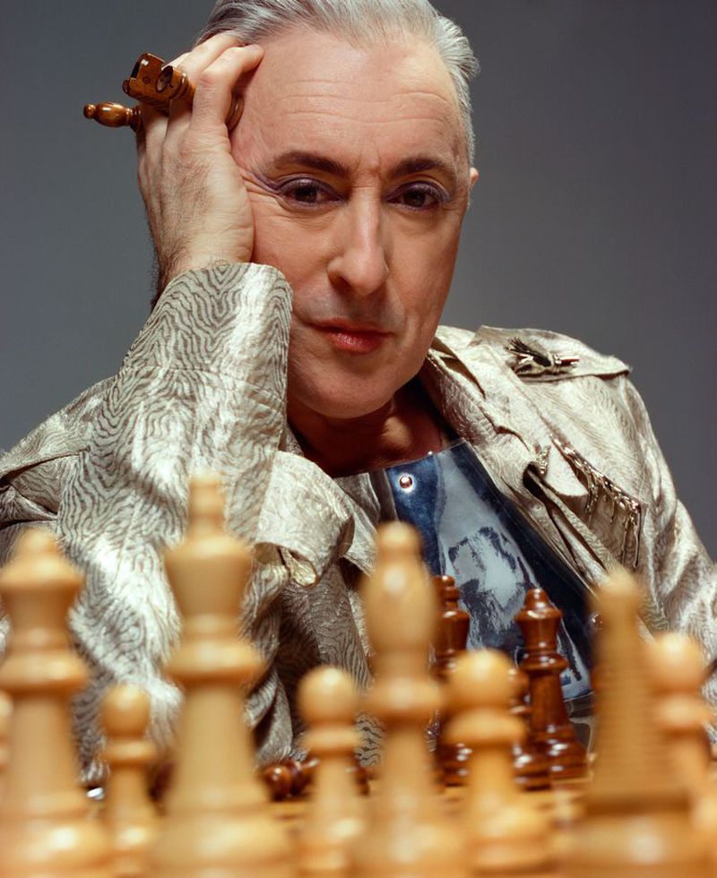 Mature adult with short grey hair poses with thoughtful expression over a chess board, wearing patterned jacket and rings. Portrait.