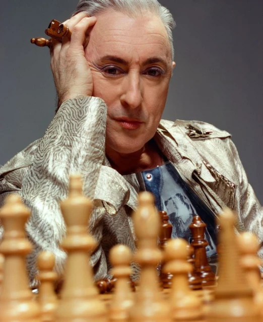 Mature adult with short grey hair poses with thoughtful expression over a chess board, wearing patterned jacket and rings. Portrait.