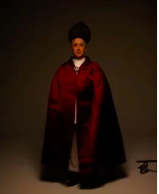 An adult male poses indoors wearing a burgundy cape and a black hat, standing in a neutral studio setting with soft, diffused lighting.