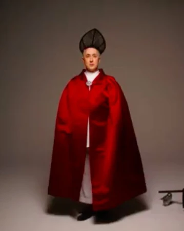 Adult male in a photo studio wearing a long, bright red cape and a black headdress, posing against a gray backdrop.