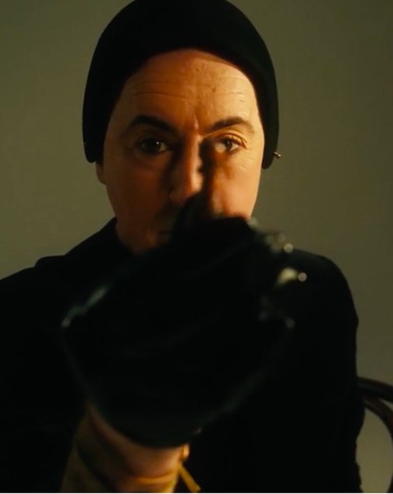 Man wearing a black beanie and turtleneck sweater holds an out-of-focus black object to the camera in a medium studio shot.