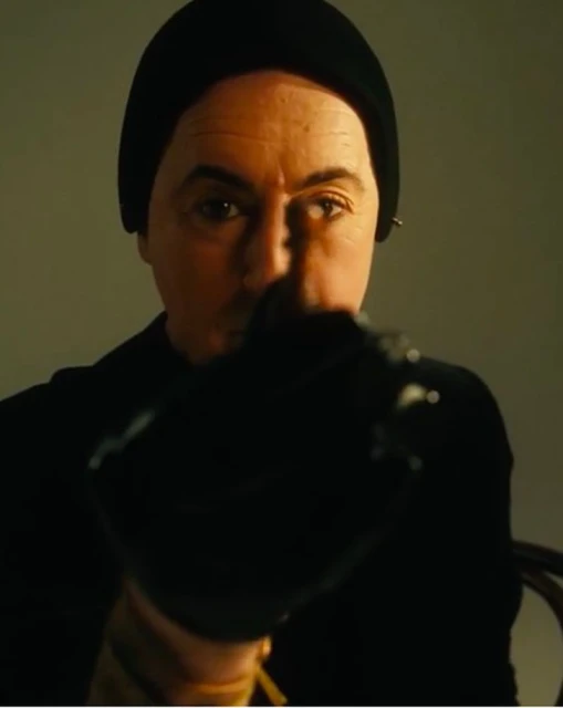 Man wearing a black beanie and turtleneck sweater holds an out-of-focus black object to the camera in a medium studio shot.