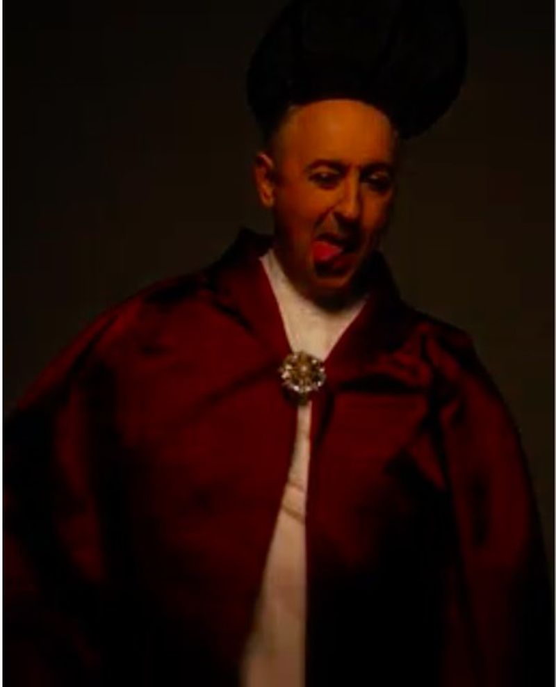 Adult male wearing a red cape with a jeweled clasp and a black hat is sticking his tongue out against a dark studio background.