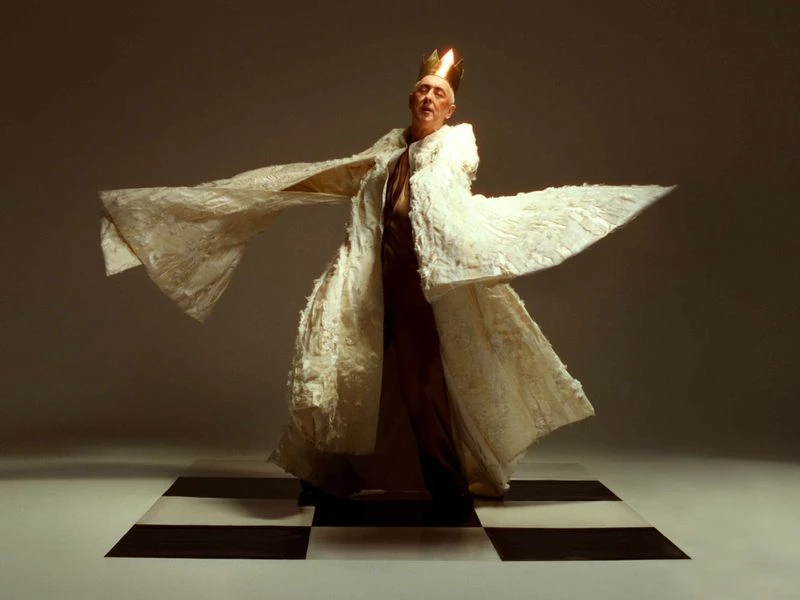 Person wearing a white textured robe and golden crown stands on a black and white checkered surface against a neutral background.