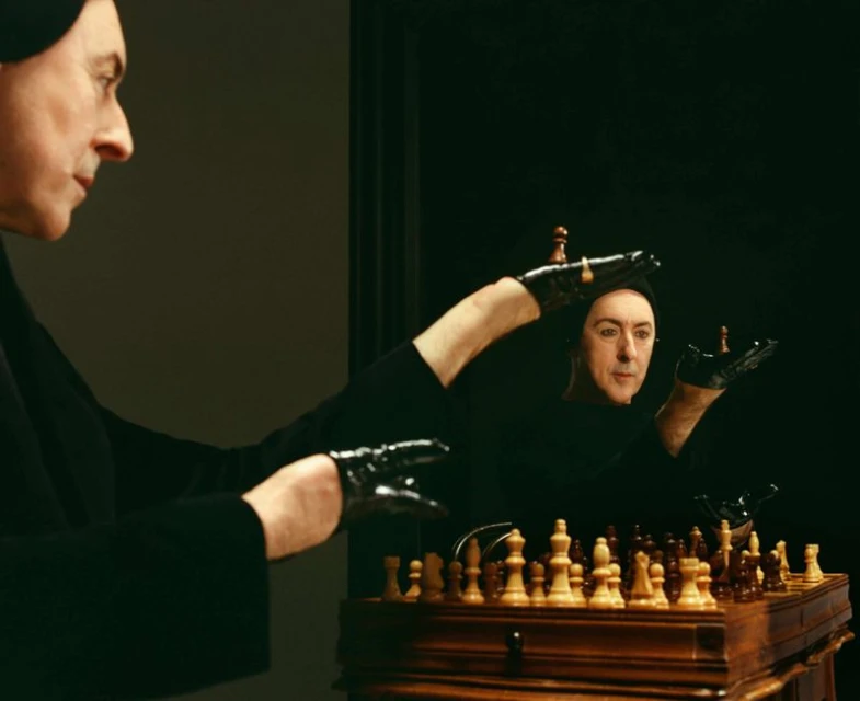 Man in black clothing plays chess with his reflection in a dark room, holding chess pieces in gloved hands, next to a wooden chessboard.