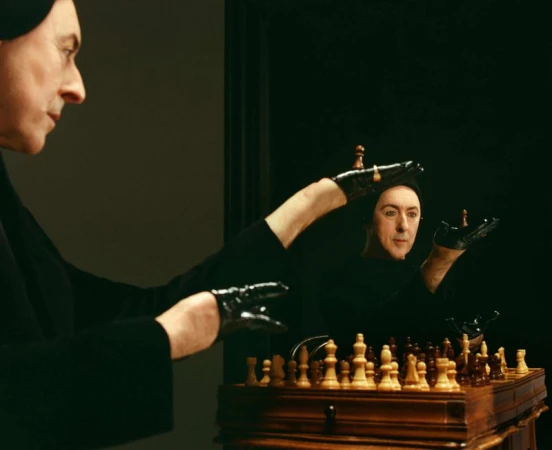 Man in black clothing plays chess with his reflection in a dark room, holding chess pieces in gloved hands, next to a wooden chessboard.