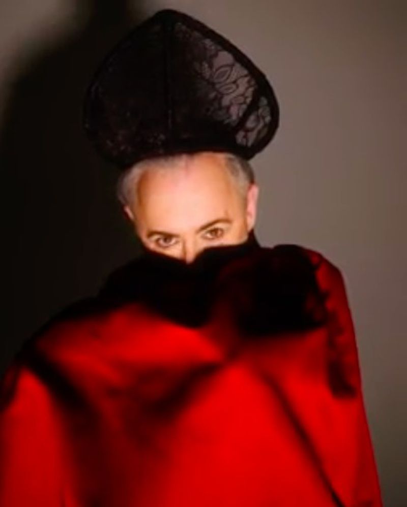 Portrait of a person with gray hair, eye contact, wearing a red garment and a black lace headdress against a gray background.
