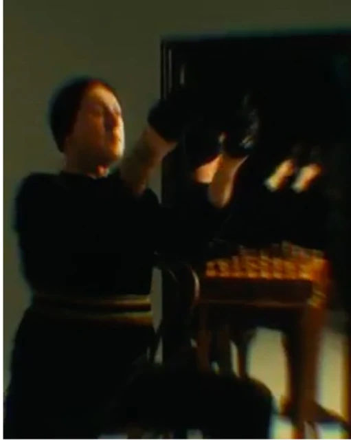 Adult male in black clothing seated at piano indoors, playing music with hands raised, captured in a dimly lit scene with soft focus.