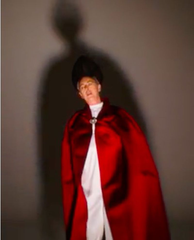 Adult in ornate red and white robe and cap standing indoors with soft lighting and a shadow in the background.