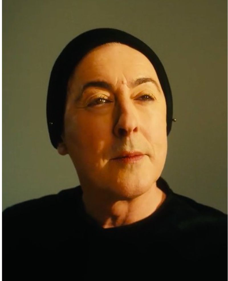 Portrait of an older woman with pale skin wearing a black head covering and top, illuminated by golden lighting against a grey backdrop.