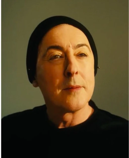Portrait of an older woman with pale skin wearing a black head covering and top, illuminated by golden lighting against a grey backdrop.