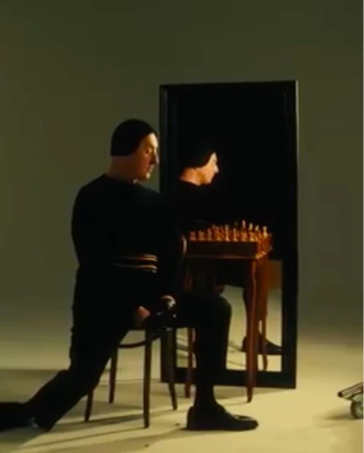 A man in black clothing playing chess. There is a reflection of the man and the chess board in a mirror behind the table.