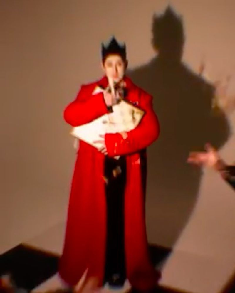 Person in a red robe and crown holds a book, finger to their lips, standing on a black and white checkerboard surface.