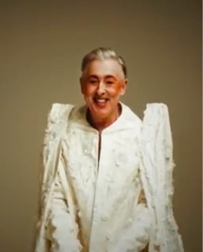 Smiling adult with white hair wearing an elaborate white costume, standing against a plain background in a medium studio shot.