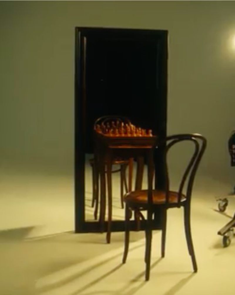 Studio shot featuring a wooden chair, a small table with a chessboard, and a large mirror in a minimalist setting with soft, diffused lighting.