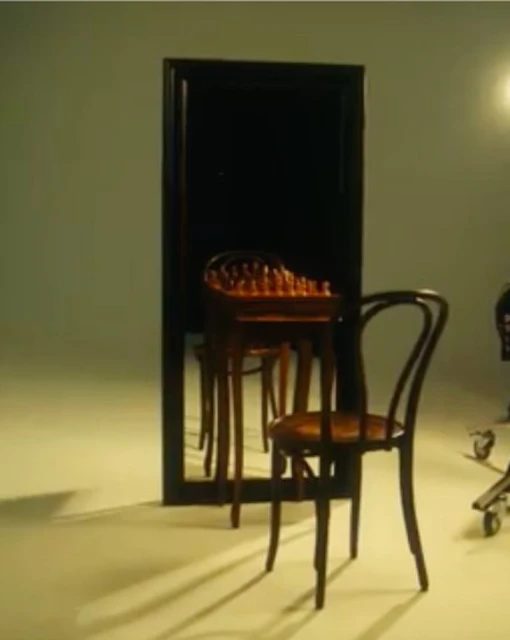 Studio shot featuring a wooden chair, a small table with a chessboard, and a large mirror in a minimalist setting with soft, diffused lighting.