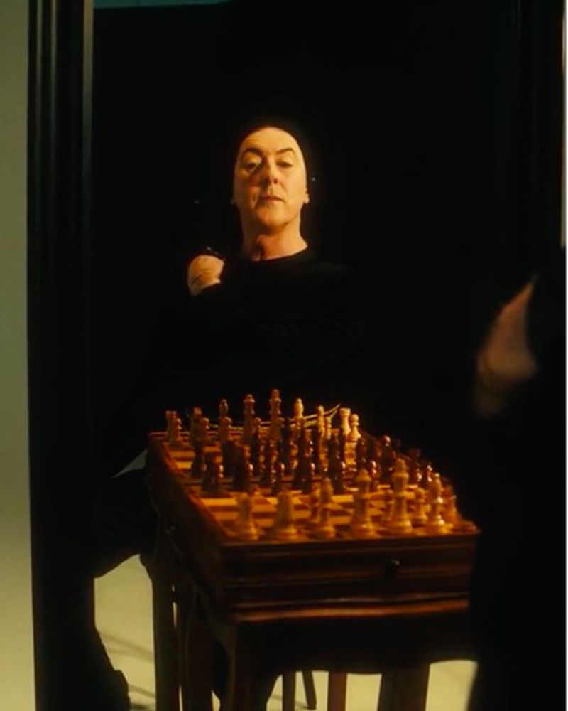An adult person in a black top plays chess on a wooden chess set. The backdrop is solid black in a dimly lit, indoor setting.