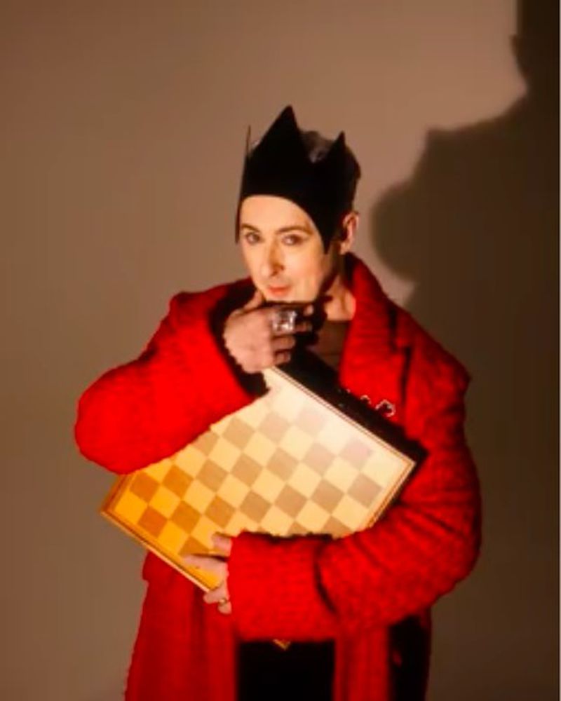 Adult male in red coat and black crown holding chessboard in indoor setting with neutral-toned background. Focused gaze, unique style.