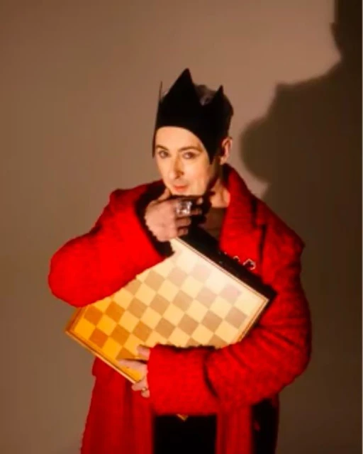 Adult male in red coat and black crown holding chessboard in indoor setting with neutral-toned background. Focused gaze, unique style.